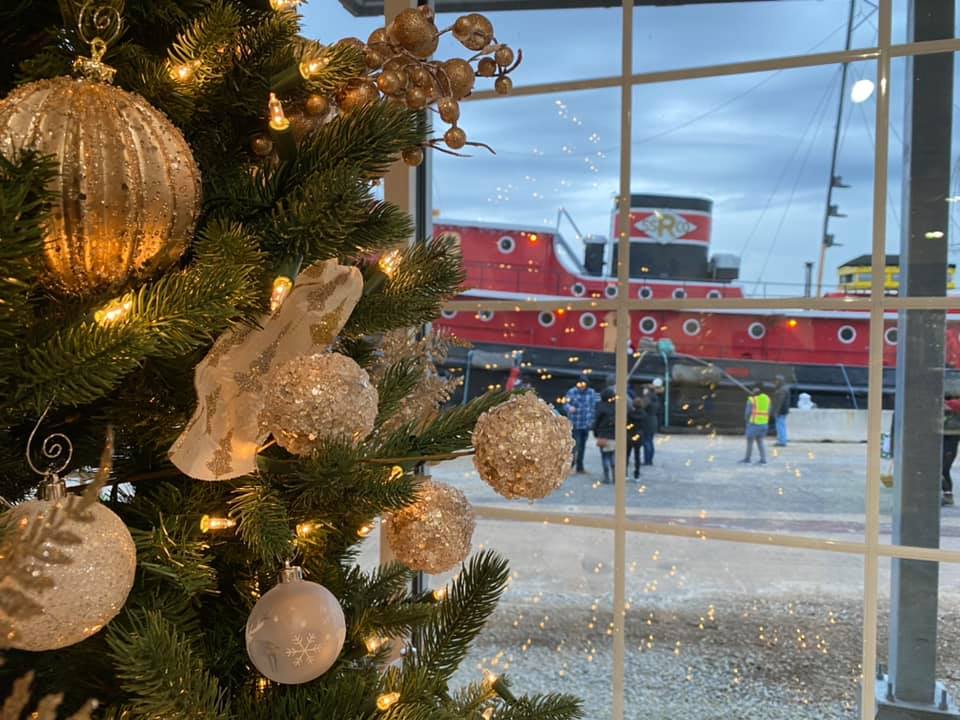 A holiday tree with golden ornaments in front of a window that shows an historic red tugboat outside.